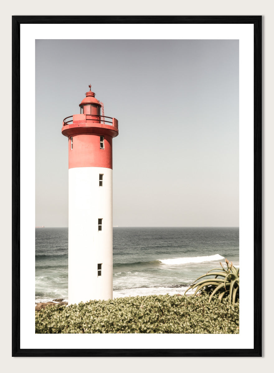 A framed photographic art print of a red and white lighthouse standing on a grassy shore with the ocean and sky in the background. Beacon by the Sea by Shot by Clint coastal photography print featuring a red-and-white lighthouse, ocean waves, pale sky, and coastal greenery.
