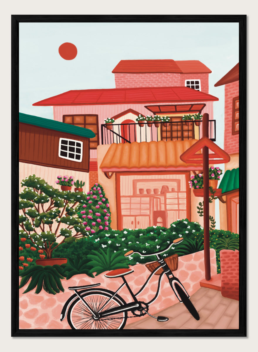 A colorful illustrated street scene with pink and peach buildings, green plants, flowers, a bicycle in the foreground, and a pale sky with a red sun. Shop Charming Village Street by Nidhi Kachhadiya, a colorful village wall art print featuring a bicycle, flower-filled homes, terracotta roofs, and warm pink and green tones.
