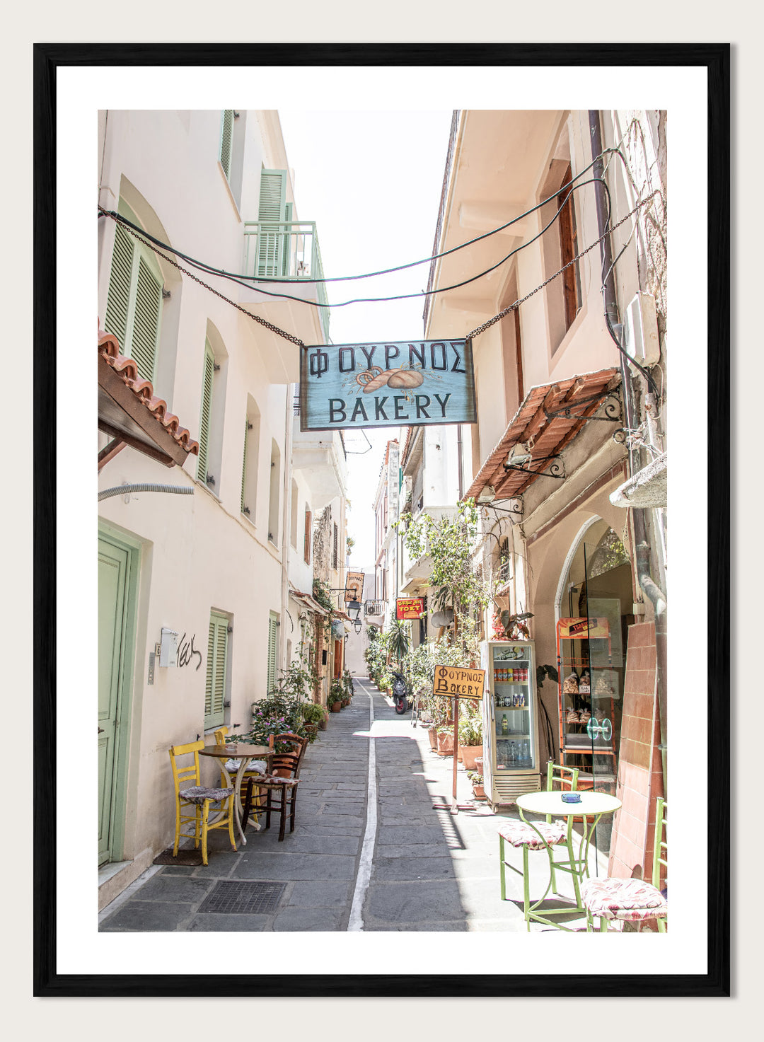  narrow street with a bakery sign in a European town. Charming Bakery Street by Henrike Schenk is a Greek street photography print featuring a Mediterranean bakery alley, pastel buildings, café seating, and travel wall art.