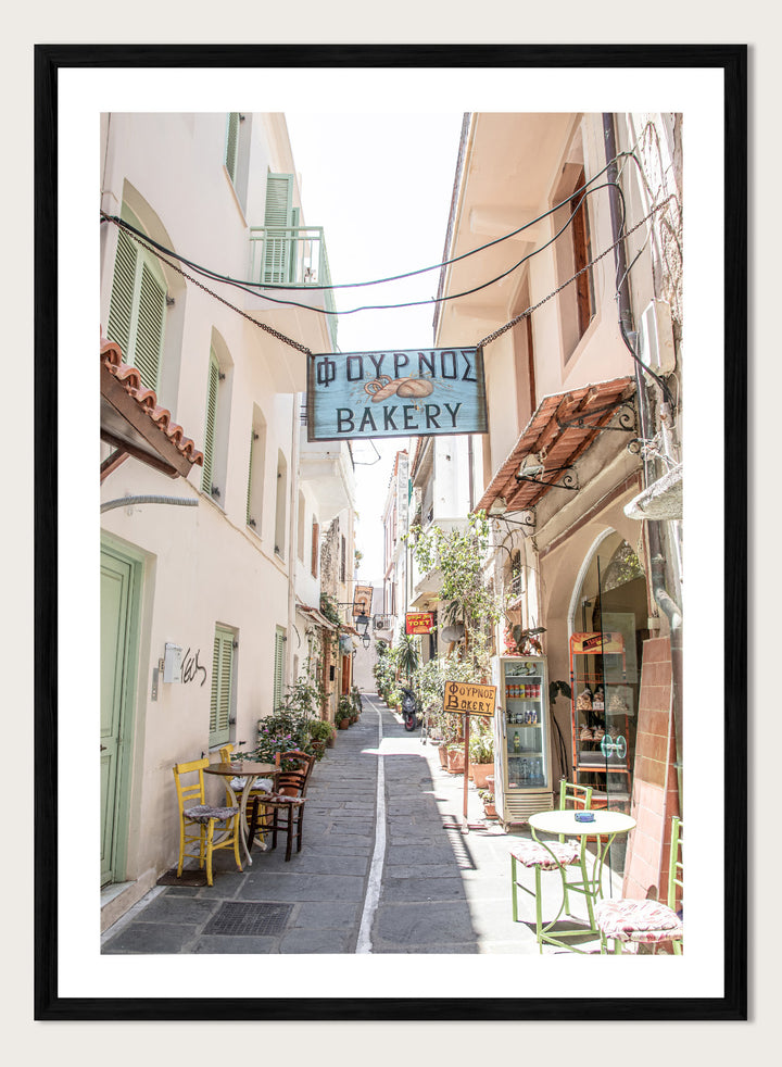  narrow street with a bakery sign in a European town. Charming Bakery Street by Henrike Schenk is a Greek street photography print featuring a Mediterranean bakery alley, pastel buildings, café seating, and travel wall art.
