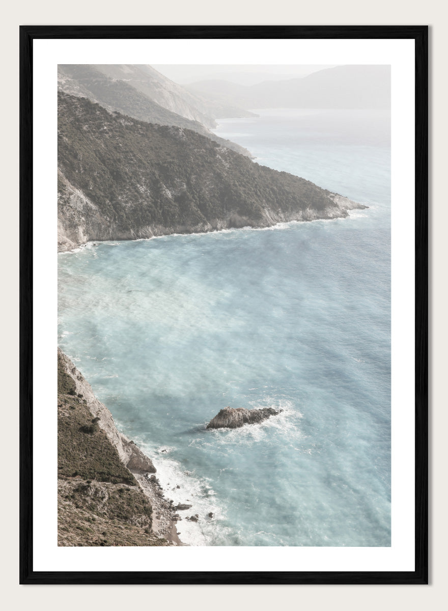 A framed photographic art print depicting a coastal landscape with a steep rocky cliff overlooking a calm, pale blue sea with a small rock formation in the foreground. Coastal Serenity by Shot by Clint coastal photography print featuring pale blue water, rugged cliffs, rocky shores, and a soft hazy horizon.