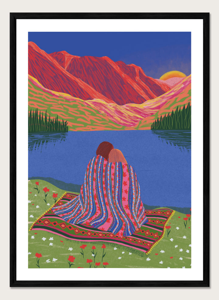 A framed art print featuring a stylized landscape illustration of two people sitting together on a patterned blanket by a blue lake, with red mountains and a yellow sun in the background. Cozy Lake Sunset art print by Ceyda Alasar features a lakeside embrace, vivid mountains, and a glowing sunset in rich, story-filled color.