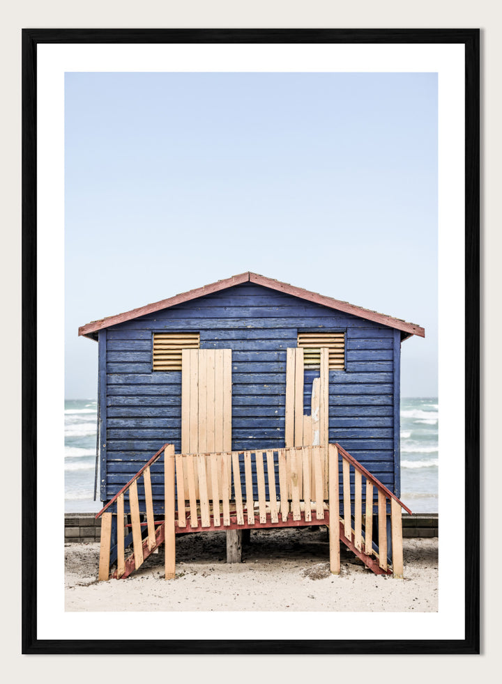 A framed photographic print of a blue wooden beach lifeguard hut on a sandy shore with the ocean and pale sky in the background. Beachside Retreat by Shot by Clint features a weathered blue beach hut and soft ocean backdrop in a calm coastal photography print.