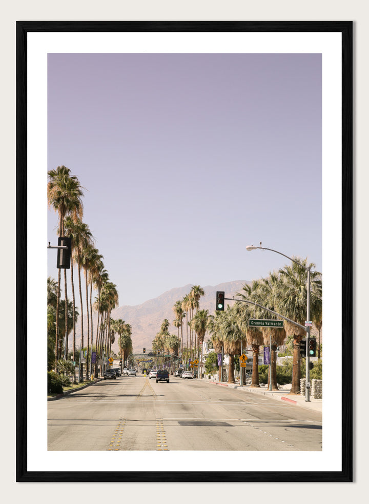 A framed art print featuring a scenic photograph of a palm-lined street in Palm Springs with mountains in the background. Palm-Lined Avenue by Shot by Clint travel photography print featuring a California road, tall palm trees, desert mountains, and soft sun-faded tones.