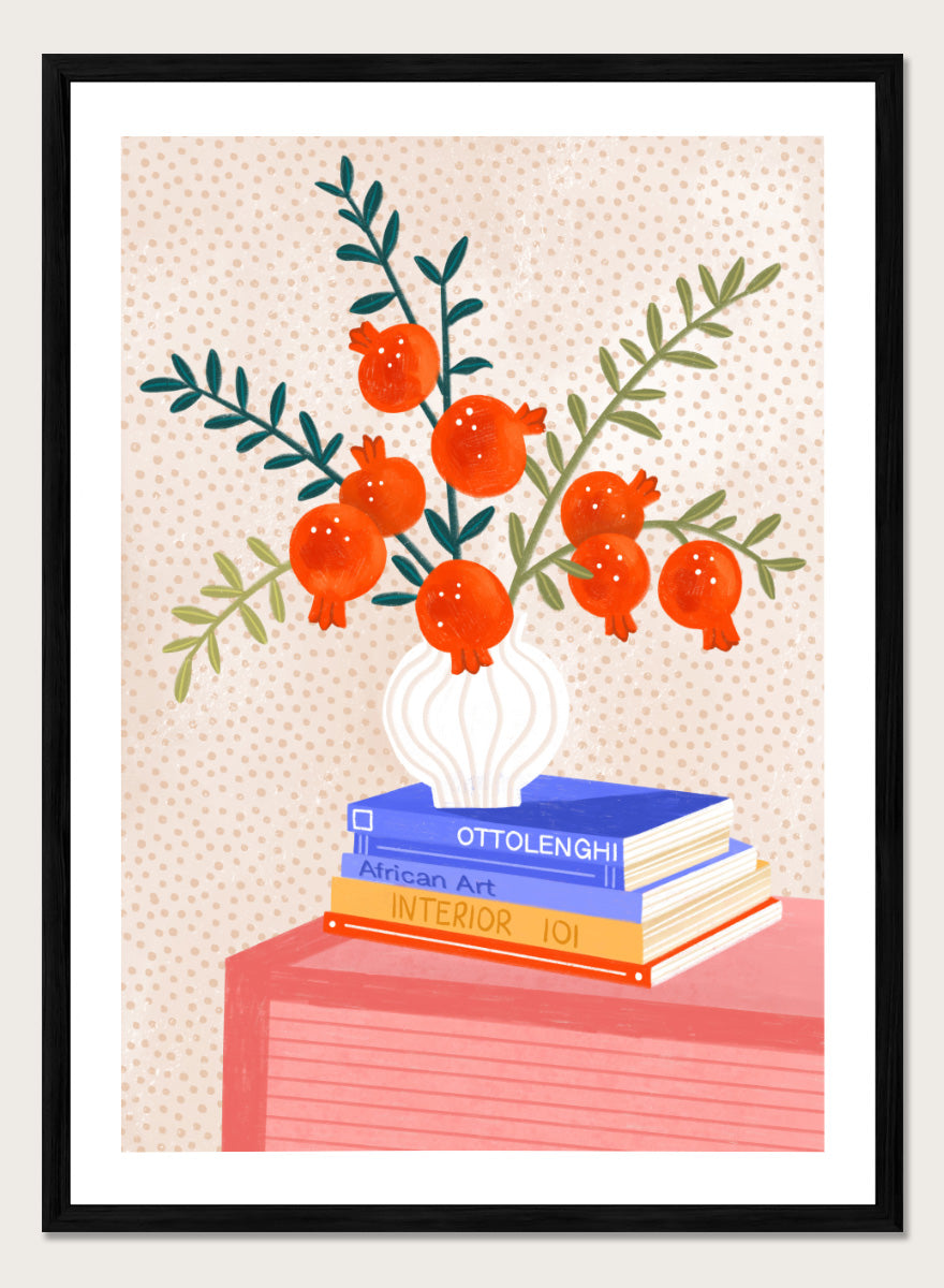 A framed art print featuring a stylized illustration of pomegranate branches in a white vase resting on a stack of books, set against a light beige dotted background. Cozy Tabletop Pomegranates by Bea Müller still life art print featuring red fruit branches, a sculptural vase, design books, and colorful interior styling.