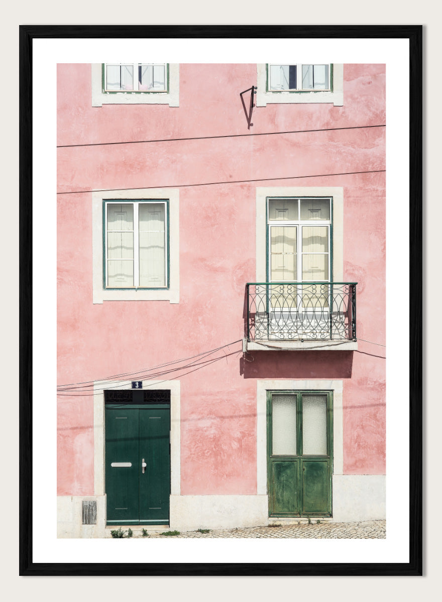 A framed art print featuring a photograph of a pink building facade with green doors, white-framed windows, and a small black metal balcony. Pink Façade Harmony by Shot by Clint architectural photography print featuring a pink exterior, green doors, white windows, and an iron balcony.