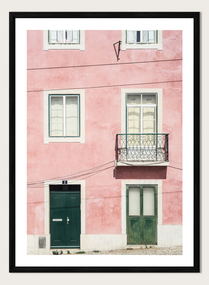 A framed art print featuring a photograph of a pink building facade with green doors, white-framed windows, and a small black metal balcony. Pink Façade Harmony by Shot by Clint architectural photography print featuring a pink exterior, green doors, white windows, and an iron balcony.