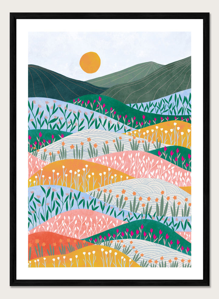 A framed art print featuring a stylized landscape illustration with rolling hills, wildflowers, mountains, and a sun in pastel and bright colors. Colorful Flower Hills by Ceyda Alasar botanical landscape art print featuring layered wildflower fields, rolling hills, mountains, and a golden sun.