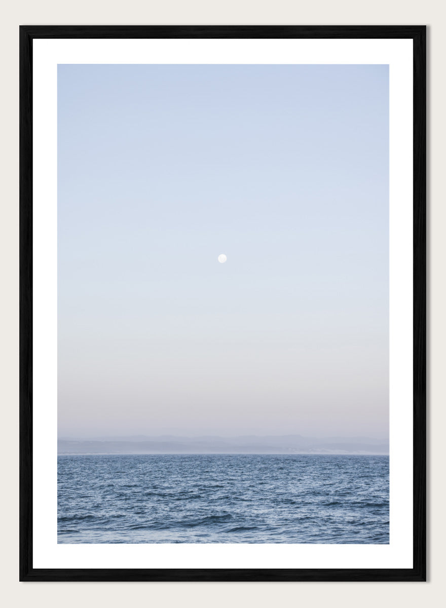 A minimalist framed art print depicting a calm blue ocean under a pale sky with a small visible moon. Moonlit Horizon by Shot by Clint coastal photography print featuring a pale moon, distant shoreline, open blue water, and soft lavender evening tones.