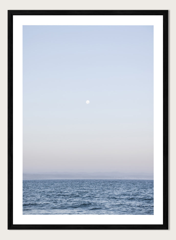A minimalist framed art print depicting a calm blue ocean under a pale sky with a small visible moon. Moonlit Horizon by Shot by Clint coastal photography print featuring a pale moon, distant shoreline, open blue water, and soft lavender evening tones.