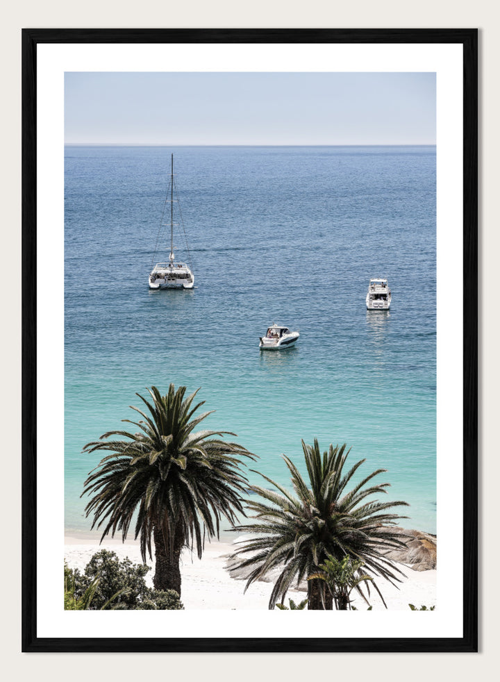 A framed coastal photograph showing a calm blue ocean with three boats, turquoise water near a white sandy shore, and two palm trees in the foreground. Tropical Serenity by Shot by Clint coastal photography print featuring turquoise water, sailboats, palm trees, and a bright tropical shoreline.