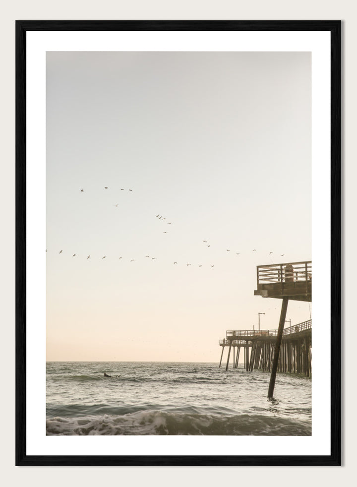 A framed photographic art print depicting a pier extending over a calm ocean under a pale sky, with birds flying across the scene. Seaside Serenity by Shot by Clint coastal photography print featuring ocean waves, a weathered pier, flying birds, and soft sunlit tones.