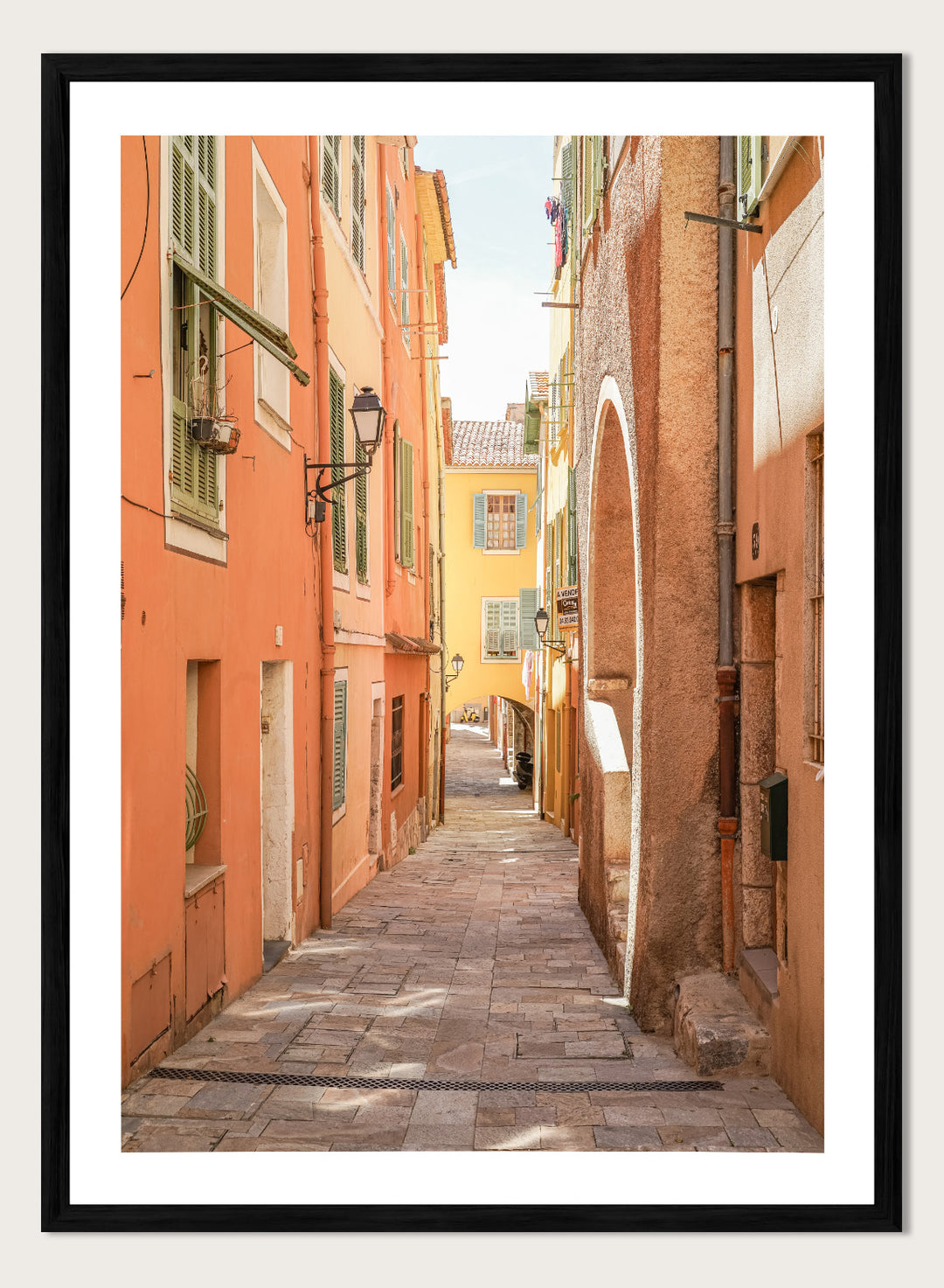 Framed photograph of a narrow street on a wall next to a window.  narrow street with colorful buildings in a European city. Vibrant European Alley by Henrike Schenk is a travel photography print featuring a Mediterranean street, cobblestone alley, and pastel European architecture.