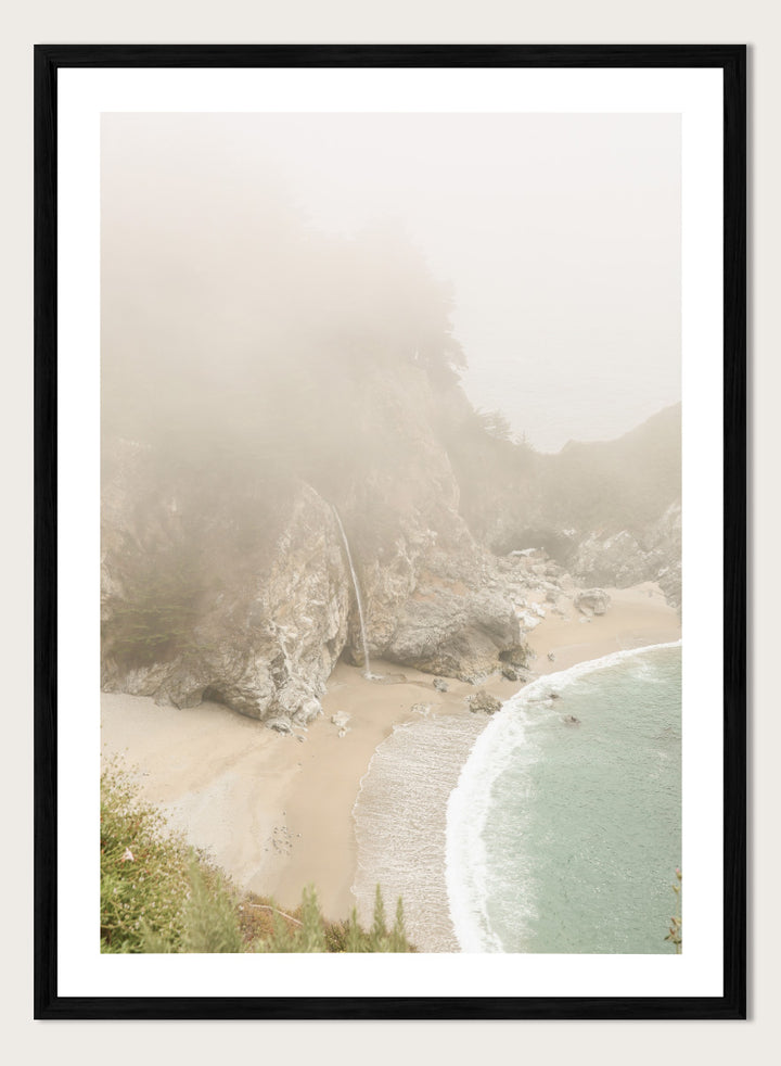 A framed photographic print of a misty coastal beach scene with a small waterfall, sandy shore, and pale blue-green ocean waves. Misty Coastal Haven by Shot by Clint coastal photography print featuring a secluded cove, rocky cliffs, pale turquoise water, and a soft veil of mist.