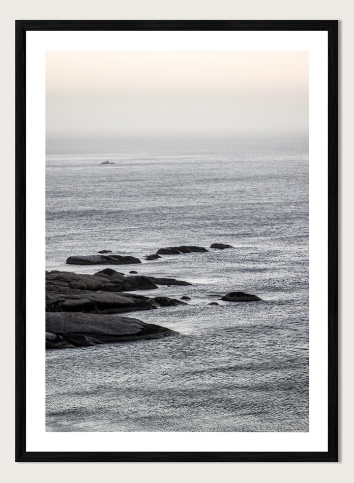 A framed black and white coastal landscape photograph featuring a calm sea, distant rocks, and a pale sky. Silver Sands by Shot by Clint coastal photography print featuring silver-blue water, dark shoreline rocks, and a soft misty horizon.