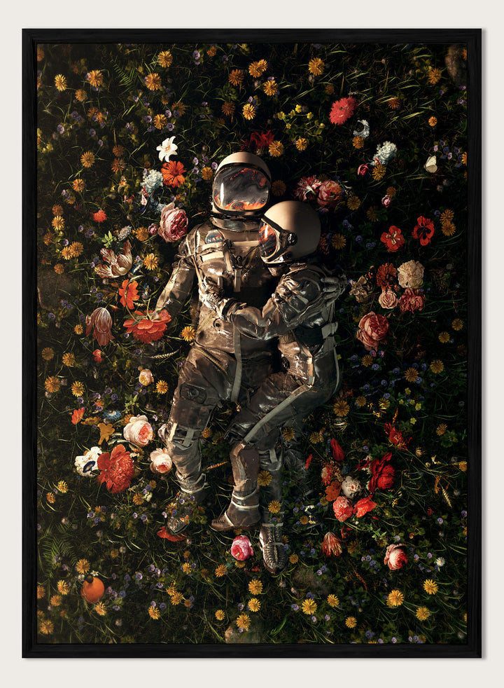 Astronaut lying on a bed of flowers with a dark background. Cosmic Couple Bloom by Nicebleed is a surreal astronaut floral art print featuring two astronauts in a field of flowers, modern aesthetic wall poster.