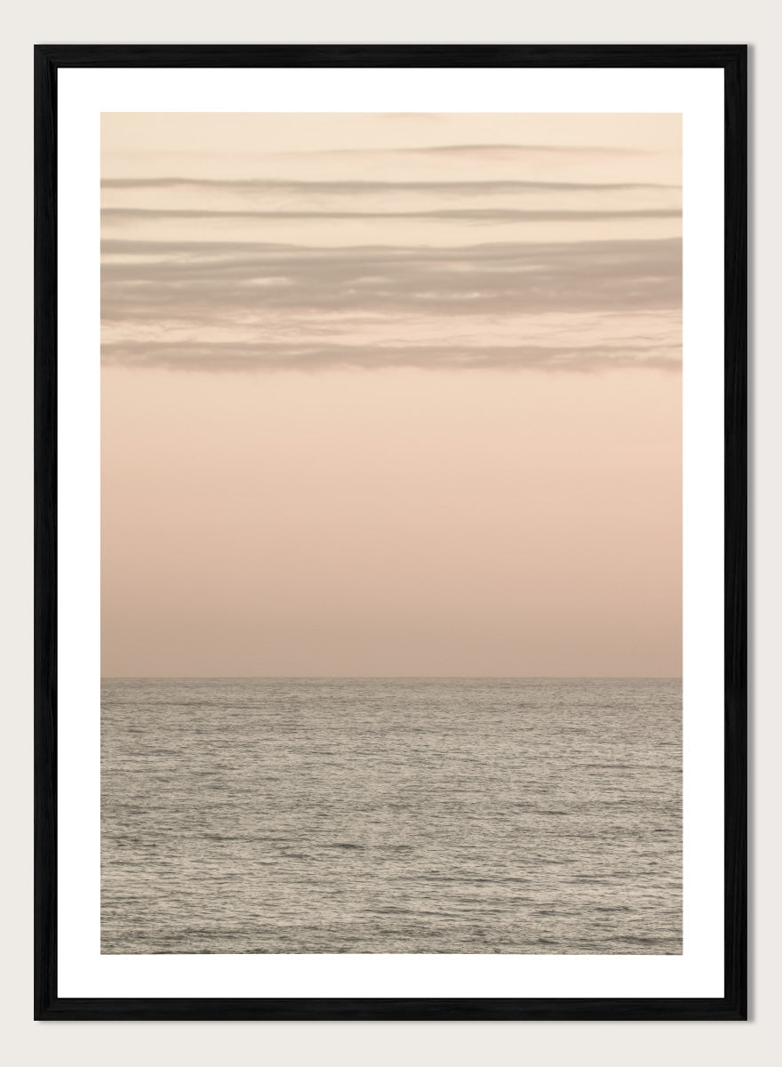 A minimalist framed art print depicting a calm seascape at dusk or dawn, with a soft peach and beige sky meeting a muted gray-blue ocean horizon. Serene Horizons by Shot by Clint features a soft peach sky and calm ocean horizon in a minimal coastal photograph with airy neutral tones.