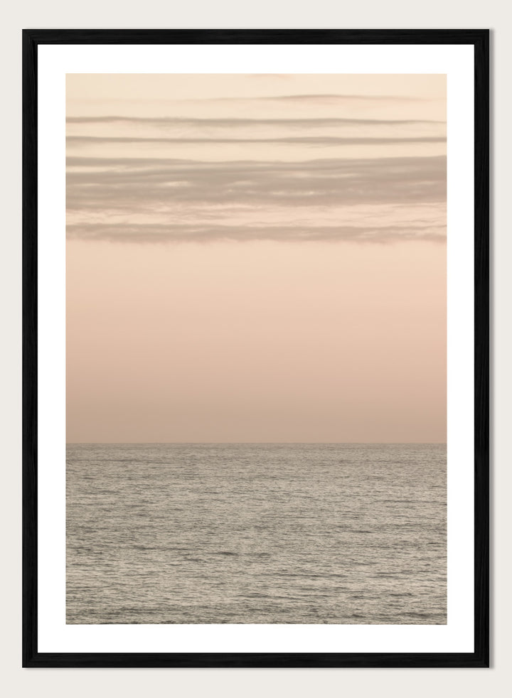 A minimalist framed art print depicting a calm seascape at dusk or dawn, with a soft peach and beige sky meeting a muted gray-blue ocean horizon. Serene Horizons by Shot by Clint features a soft peach sky and calm ocean horizon in a minimal coastal photograph with airy neutral tones.