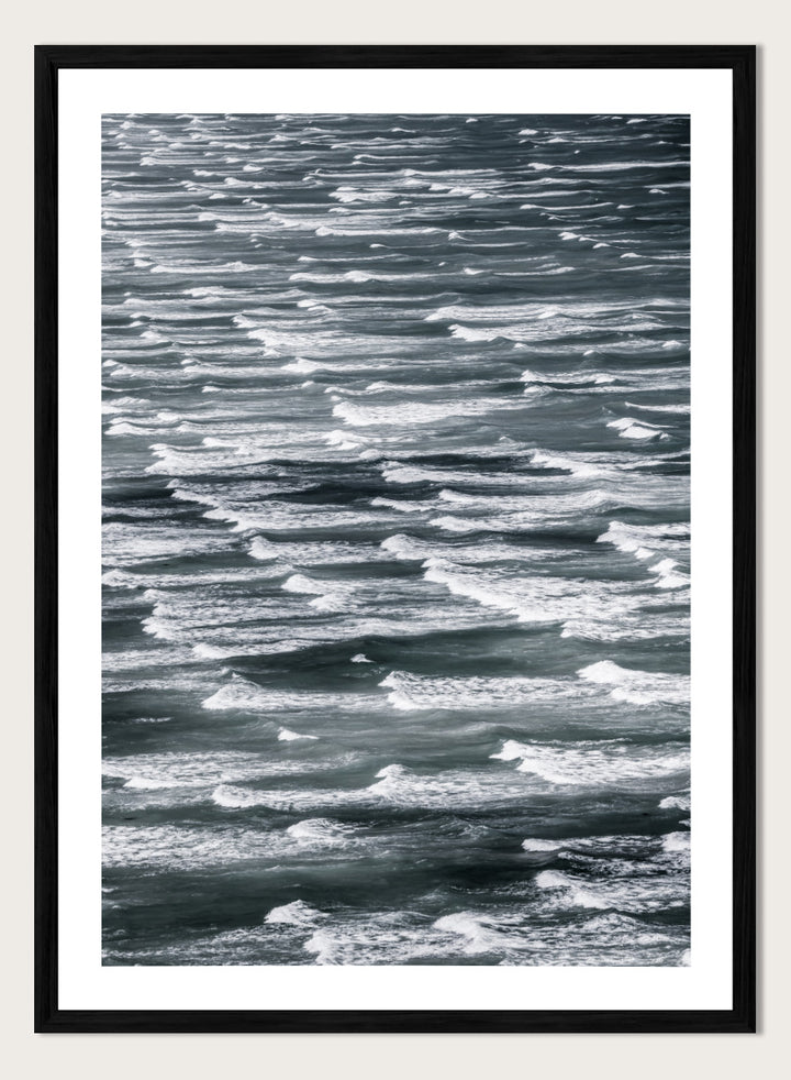 A framed black and white photographic print depicting a close-up view of ocean waves with white foam crests against dark water. Waves in Rhythm by Shot by Clint ocean photography print featuring layered blue-grey waves, white foam, and powerful natural movement across the sea.