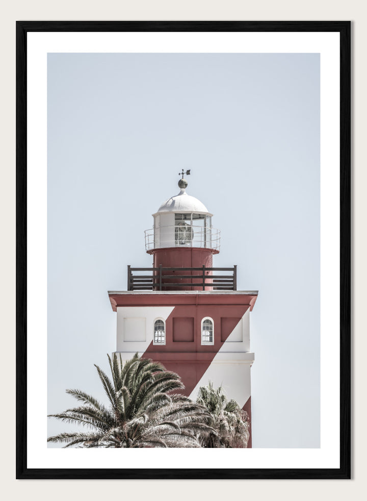 A framed art print featuring a red and white lighthouse with palm trees at the bottom, set against a pale sky background. Beacon of Solitude by Shot by Clint lighthouse photography print featuring red-and-white architecture, palm trees, arched windows, and a pale coastal sky.