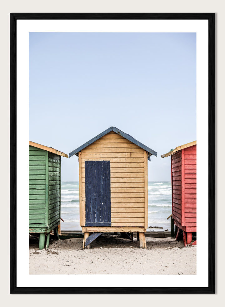 A framed photographic print of three colorful wooden beach huts on a sandy beach, with the central hut in natural wood and a dark blue door, flanked by green and red huts under a pale blue sky. Colorful Beach Huts by Shot by Clint coastal photography print featuring green, natural wood, and red huts with weathered texture and ocean views.