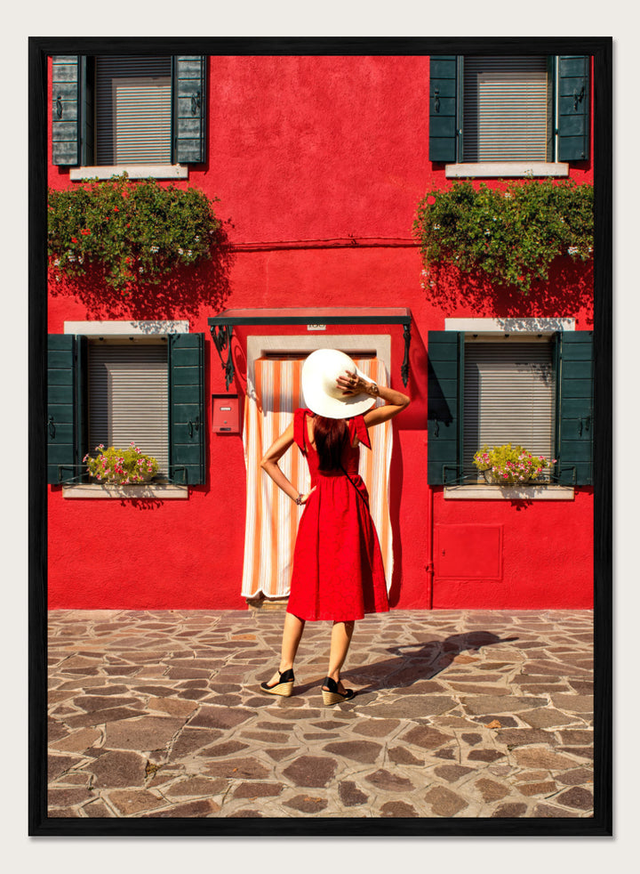 Woman in a red dress and white hat standing in front of a red wall with green shutters. Red Art Print by Anette Ohlendorf featuring a woman in a red dress standing against a red building. Officially licensed travel photography with strong color and symmetry.