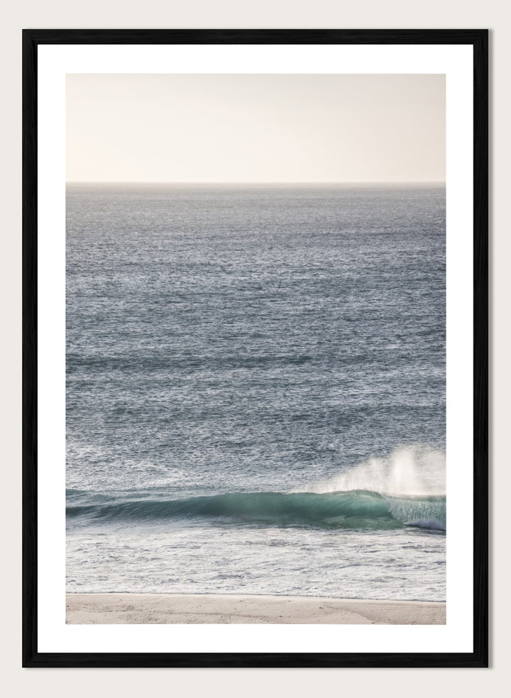 A framed photographic art print of an ocean scene, featuring a gentle wave breaking on a sandy beach under a pale sky. Tranquil Ocean Scene by Shot by Clint ocean photography print with a rolling wave, soft sea spray, and airy blue coastal tones.