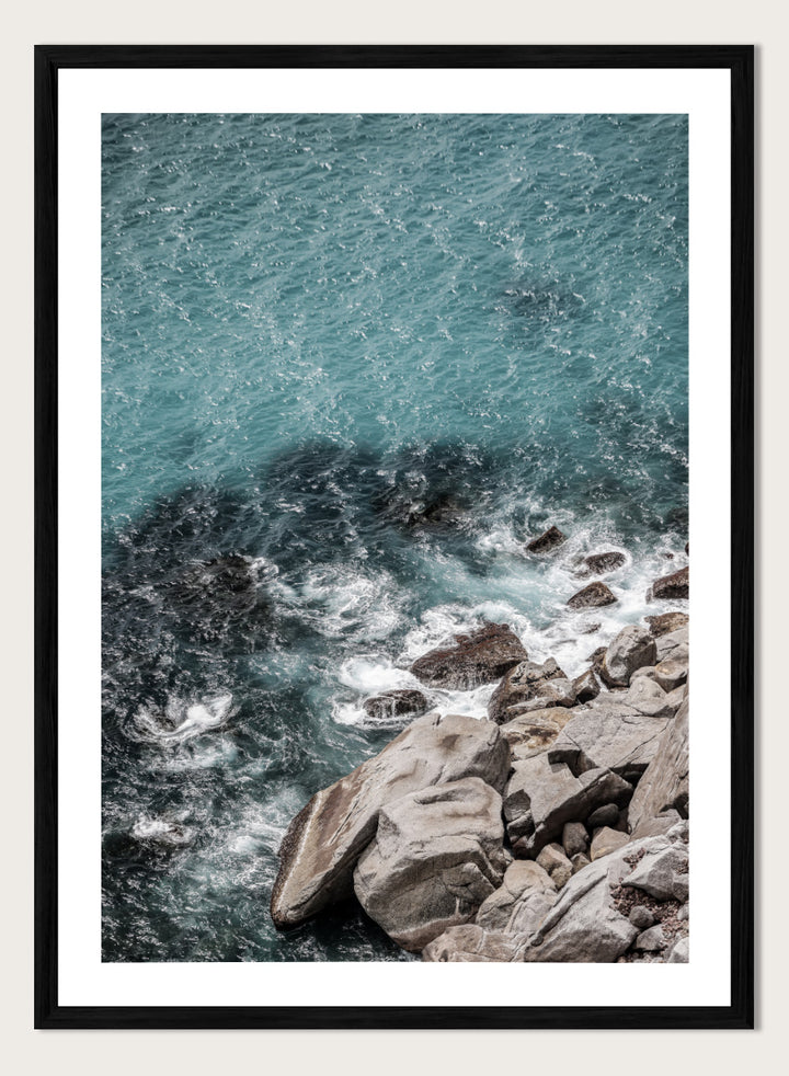A framed photographic art print depicting a top-down view of turquoise ocean water washing over dark rocks and a rocky shoreline. Ocean’s Edge Symphony by Shot by Clint aerial ocean photography print featuring turquoise water, white surf, coastal texture, and soft seafoam tones.