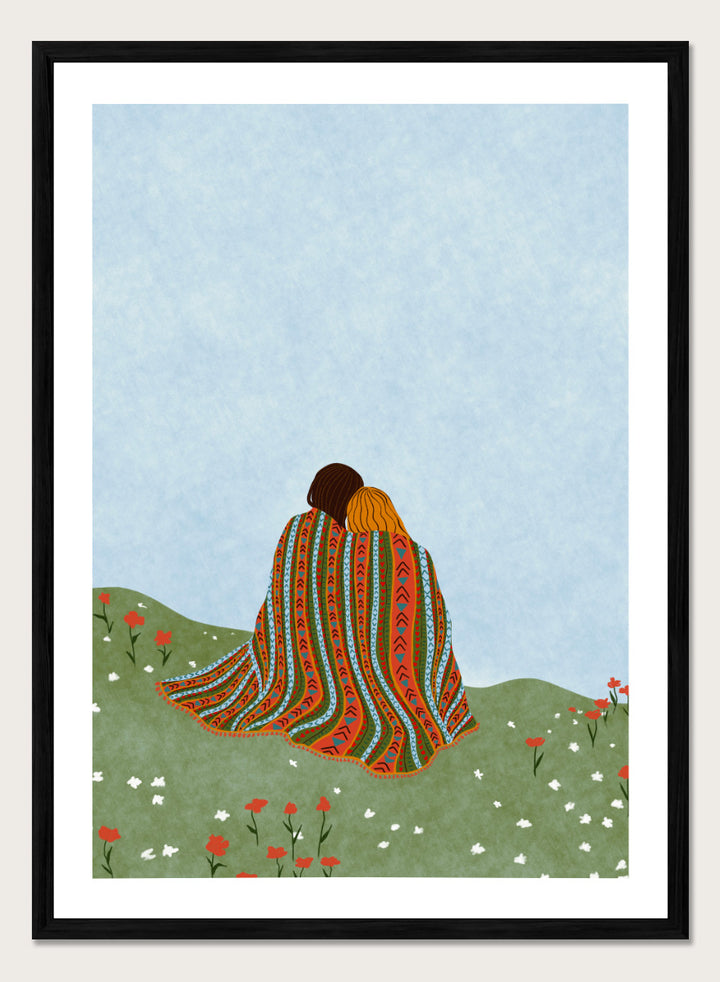 A framed art print featuring an illustration of two people sitting on a grassy hill, wrapped in a vibrant, patterned blanket under a light blue sky. Cozy Hillside Embrace by Ceyda Alasar art print featuring two people wrapped in a patterned blanket among green hills and wildflowers.