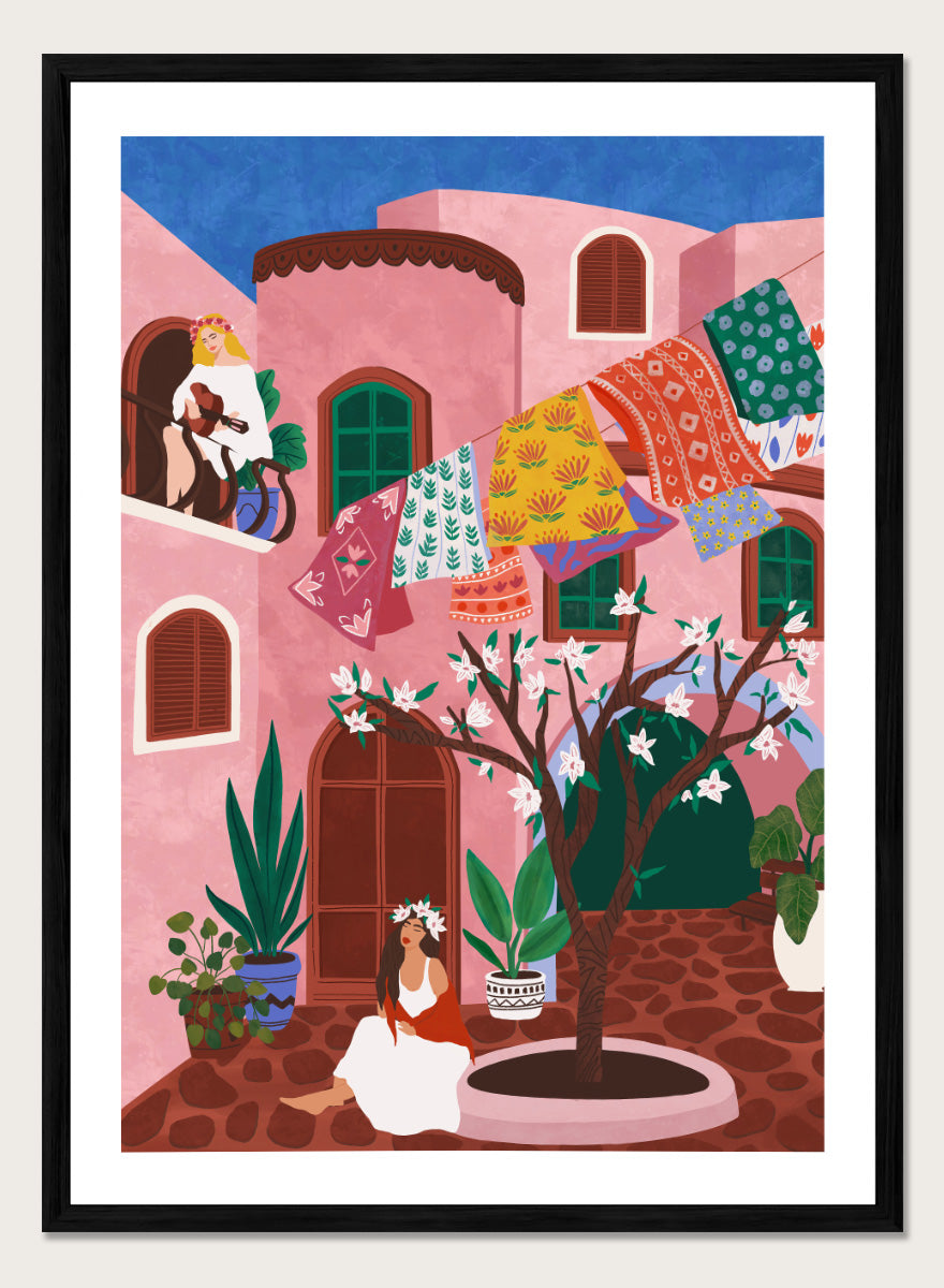 A framed art print featuring a colorful Mediterranean-style illustration of a pink courtyard with a woman sitting under a flowering tree, laundry hanging in the background, and a blue sky above. Pink Village Courtyard by Ceyda Alasar art print featuring pink architecture, patterned rugs, flowering trees, potted plants, and relaxed courtyard life.