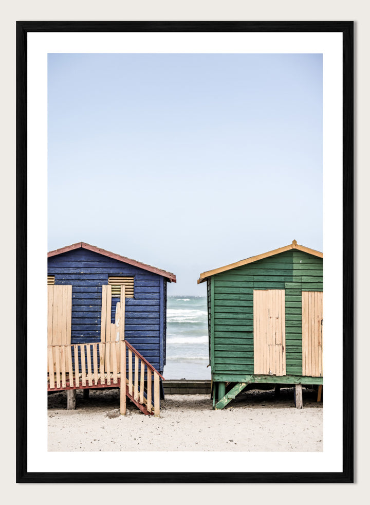 A framed photographic print of two wooden beach huts on a sandy beach under a pale blue sky, with one hut in dark blue and the other in green, both with light wood shutters and doors. Coastal Huts Harmony by Shot by Clint coastal photography print featuring blue and green beach huts, sandy ground, weathered wood, and ocean views.