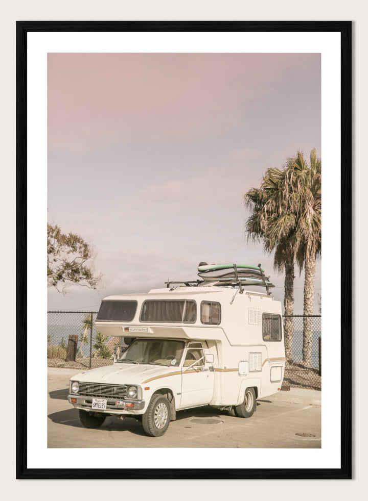 A vintage white camper truck parked outdoors near palm trees and a fence, with a surfboard strapped to its roof rack. Vintage Camper Parked by Shot by Clint travel photography print featuring a retro camper, surfboards, palm trees, and sun-faded coastal tones.
