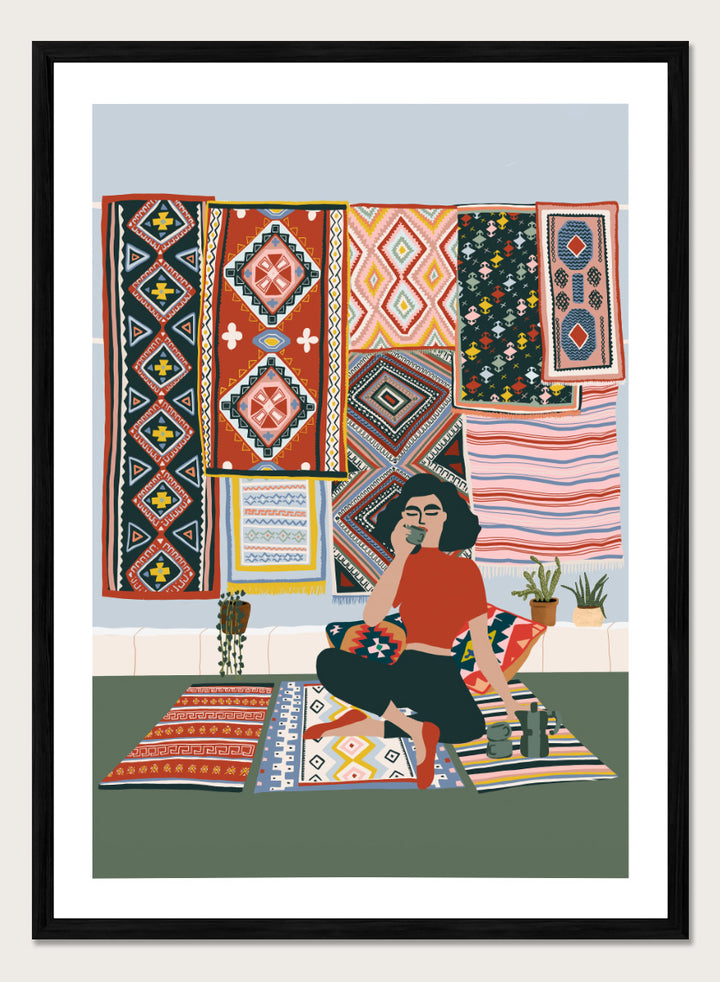 A framed art print featuring a stylized illustration of a woman sitting cross-legged on a patterned rug, surrounded by colorful geometric textiles hanging on a wall. Bohemian Coffee Morning by Ceyda Alasar interior art print featuring patterned rugs, floor cushions, potted plants, coffee, and colorful collected decor.