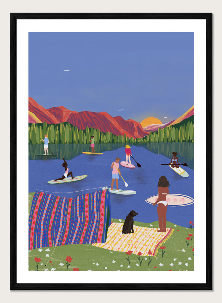 A framed art print featuring a colorful, stylized illustration of people paddleboarding and relaxing on a lake with mountains in the background. Paddleboard Lake Life by Ceyda Alasar outdoor art print featuring paddleboarders, mountain scenery, wildflowers, picnic blankets, and a lakeside dog.