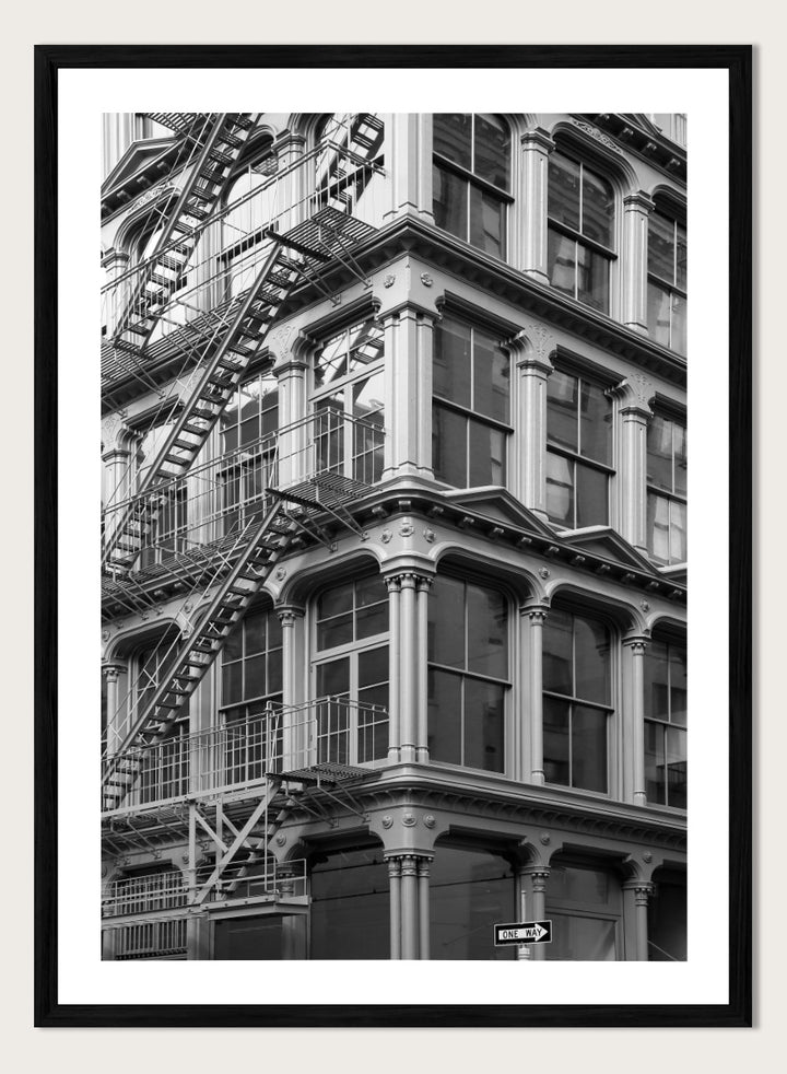 Black and white architectural photograph of a corner building facade with ornate windows and external fire escapes. Urban Geometry by Shot by Clint black-and-white architectural photography print featuring fire escapes, tall windows, ornate columns, and layered city detail.