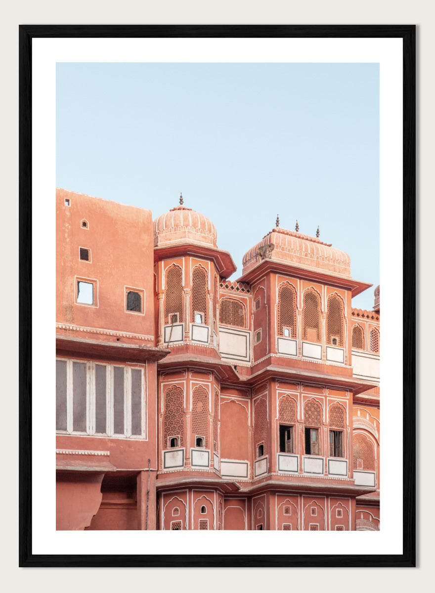 A framed art print featuring a photograph of the iconic pink sandstone Rajasthani palace, Udaipur, with white architectural details under a clear light blue sky. Terracotta Splendor by Shot by Clint architectural photography print featuring ornate palace windows, domed balconies, carved details, and warm coral tones.