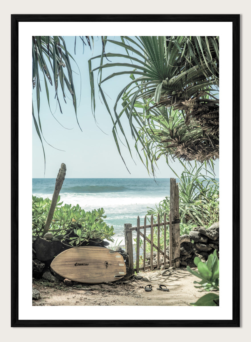 A framed beach scene art print featuring a wooden surfboard leaning against a rustic gate, with palm leaves framing a view of the ocean under a light blue sky. Coastal Serenity by Shot by Clint coastal art print featuring a surfboard, tropical plants, rustic gate, sandy path, and ocean waves.