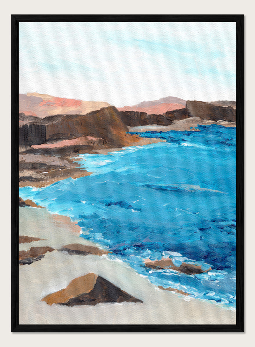 A framed landscape painting depicting a coastal scene with bright blue ocean water, sandy shore, dark rocky cliffs, and distant hills under a pale sky. Shop Rocky Coastal Waters by Nikita Jariwala, a coastal wall art print poster with blue ocean water, rocky cliffs, and a peaceful seaside view.