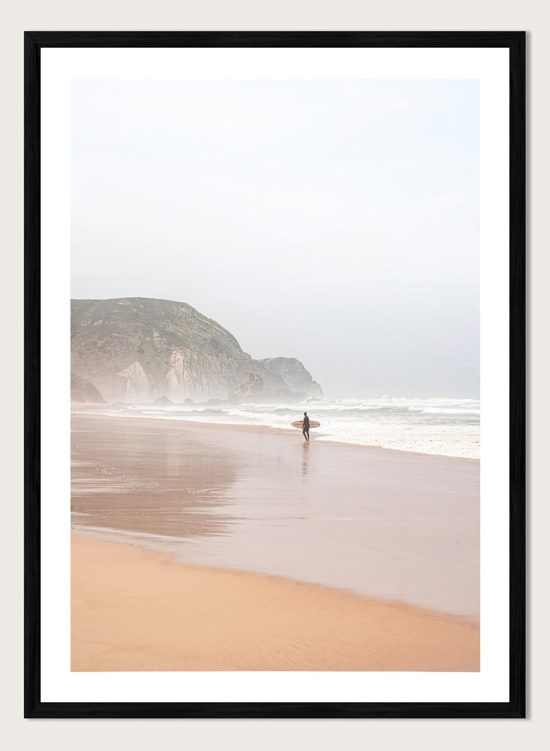Person walking along a beach with mountains in the background. Misty Morning Surfer by Henrike Schenk captures a surfer walking along a misty beach with ocean waves, coastal scenery, and photography wall art.