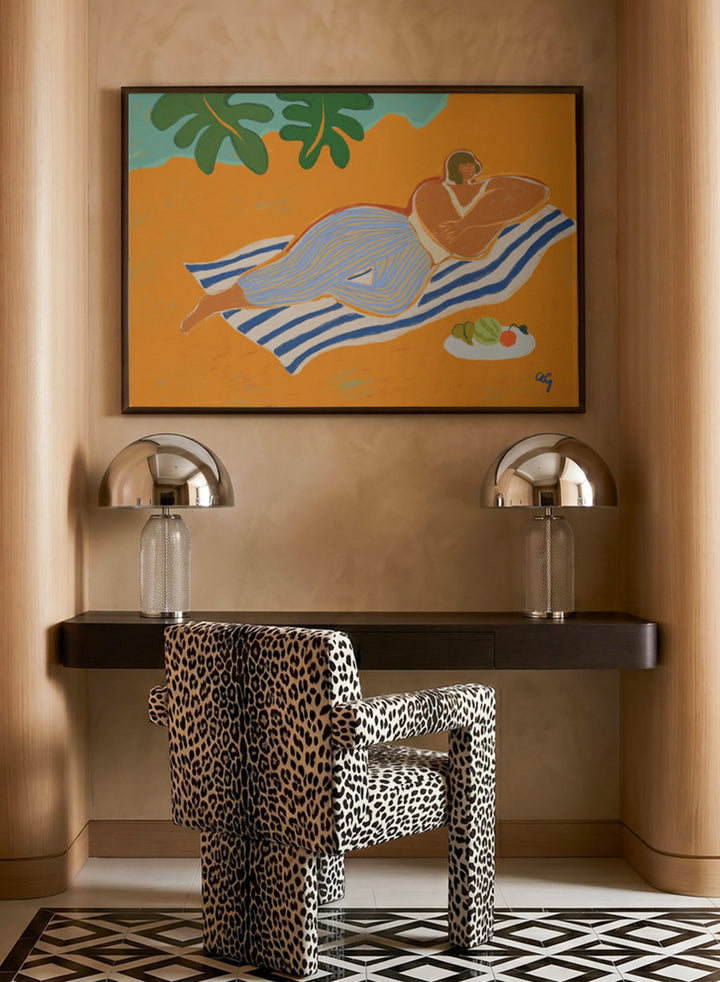 Framed artwork of a woman lounging on a towel with a plate of fruit against an orange background. A Day at the Beach by Arty Guava is a colorful coastal art print featuring a relaxed beach scene, tropical foliage, bold color, and contemporary illustration.